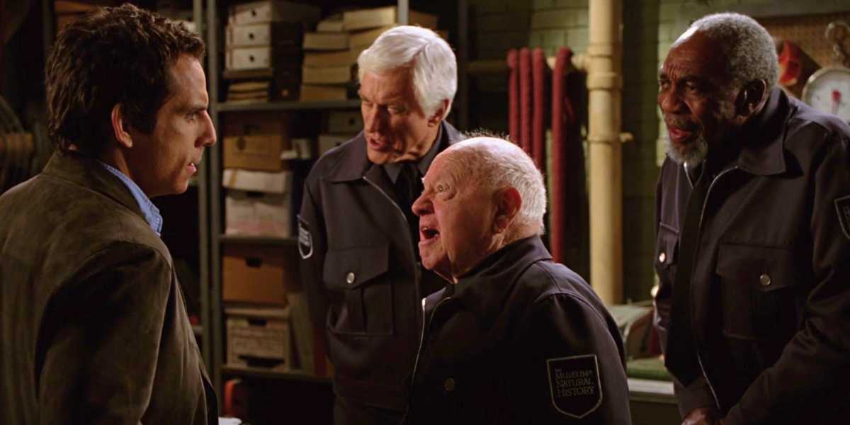A man in a brown jacket converses with three older men wearing security guard uniforms in what appears to be a storage or maintenance room. Shelves with various items and equipment can be seen in the background. The men are engaged in a lively discussion.