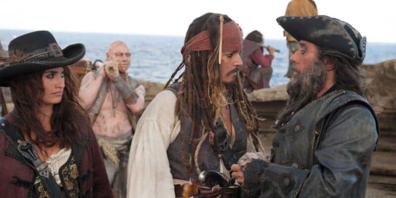 Three individuals dressed as pirates, reminiscent of Pirates of the Caribbean, are engaged in a conversation on the deck of a ship. The central figure, wearing a red bandana and beads, faces an older man in a black hat. A woman in a wide-brimmed hat stands to the left, while a rugged sailor is visible in the background.