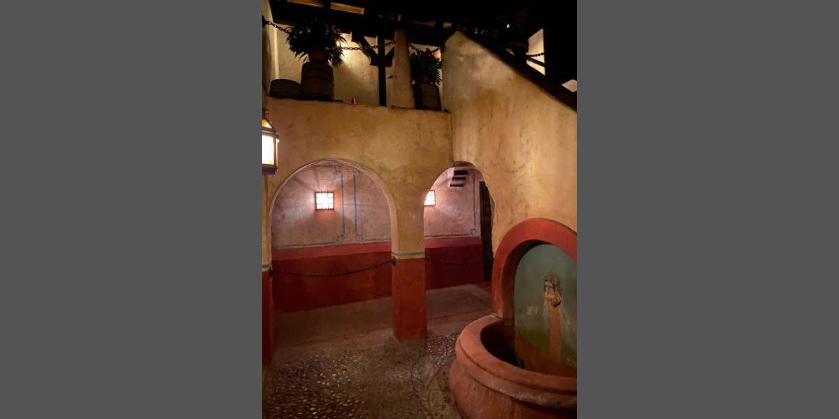 A dimly lit interior features rustic architecture with yellowish-brown walls, reminiscent of Disney World's Pirates of the Caribbean ride. Two archways, wooden ceiling beams, and a few potted plants can be seen. A well with a painted figure and a lantern hanging from the ceiling add to the atmospheric setting.