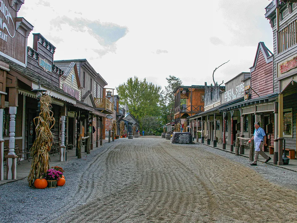 A dirt street in a Western-themed town lined with wooden buildings on both sides. The buildings have various signs, including "General Store." Decorative pumpkins and cornstalks are placed near some buildings. A few people are visible in the scene.