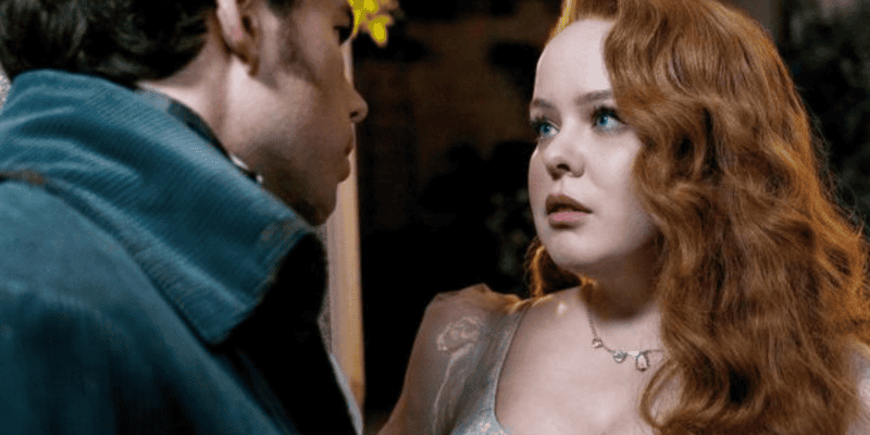 A woman with long, wavy red hair is wearing a light-colored dress and a delicate necklace. She has a concerned expression and is looking at a man with dark hair wearing a dark coat, who has his back partially to the camera. The scene appears to be dramatic.