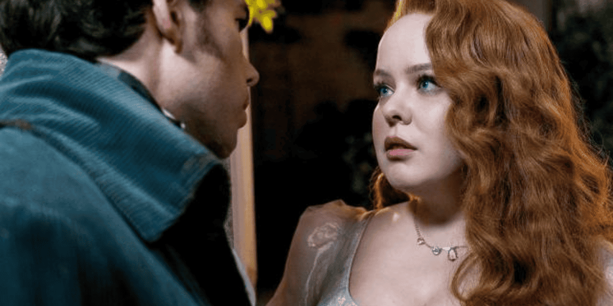 A woman with long, wavy red hair is wearing a light-colored dress and a delicate necklace. She has a concerned expression and is looking at a man with dark hair wearing a dark coat, who has his back partially to the camera. The scene appears to be dramatic.