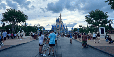 Visitors walk down a broad pathway towards a grand, iconic castle with blue and gold accents. The sky is partly cloudy, and trees line the pathway. People are taking photos, chatting, and enjoying the theme park atmosphere.