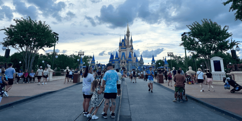 Visitors walk down a broad pathway towards a grand, iconic castle with blue and gold accents. The sky is partly cloudy, and trees line the pathway. People are taking photos, chatting, and enjoying the theme park atmosphere.