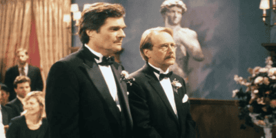 Two men in tuxedos stand side by side in a formal setting, possibly a wedding, with a statue of David in the background. One man has dark hair and the other has light brown hair and glasses. Guests are seated in the background, and there are floral arrangements nearby.