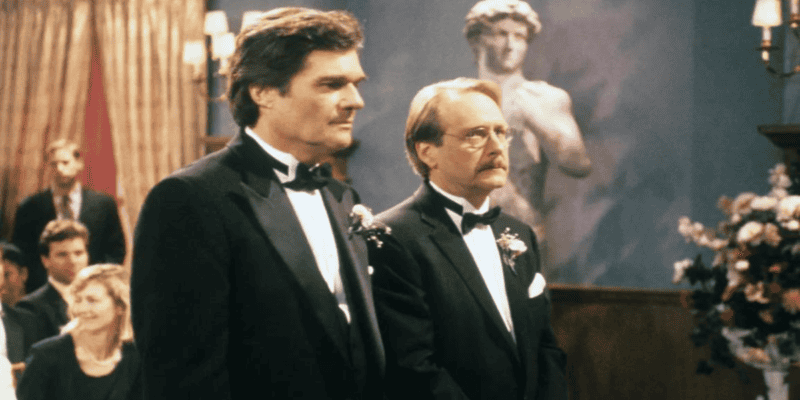 Two men in tuxedos stand side by side in a formal setting, possibly a wedding, with a statue of David in the background. One man has dark hair and the other has light brown hair and glasses. Guests are seated in the background, and there are floral arrangements nearby.