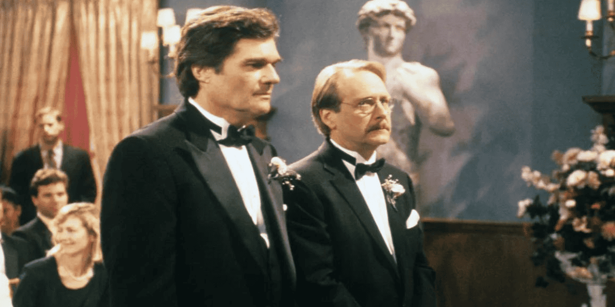 Two men in tuxedos stand side by side in a formal setting, possibly a wedding, with a statue of David in the background. One man has dark hair and the other has light brown hair and glasses. Guests are seated in the background, and there are floral arrangements nearby.