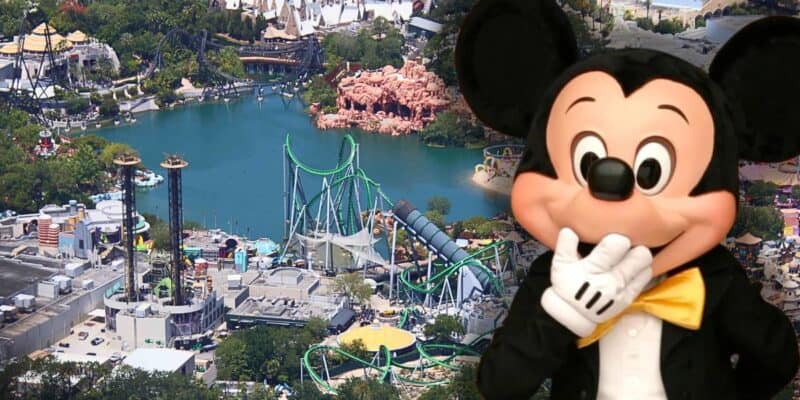An aerial view of an amusement park featuring various rides and attractions around a lake, including roller coasters and themed buildings. In the foreground, a large costumed character resembling Universal's famous Despicable Me minion is playfully holding his gloved hand to his face.