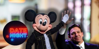 A person dressed as Mickey Mouse and Ron DeSantis in a suit raise their hands together in a celebratory gesture. On the left side of the image, there is a neon sign that reads "DATE NIGHT" with small star accents. The background is blurred, depicting an indoor setting.