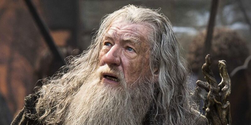 Sir Ian McKellen, dressed in old-fashioned, rugged clothing, looks upward with a surprised or concerned expression. He holds a twisted wooden staff in his right hand. The background is blurred, suggesting an outdoor or rustic setting, reminiscent of Sir Ian McKellen's iconic roles.