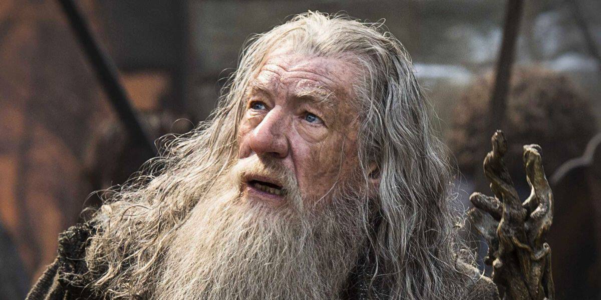Sir Ian McKellen, dressed in old-fashioned, rugged clothing, looks upward with a surprised or concerned expression. He holds a twisted wooden staff in his right hand. The background is blurred, suggesting an outdoor or rustic setting, reminiscent of Sir Ian McKellen's iconic roles.