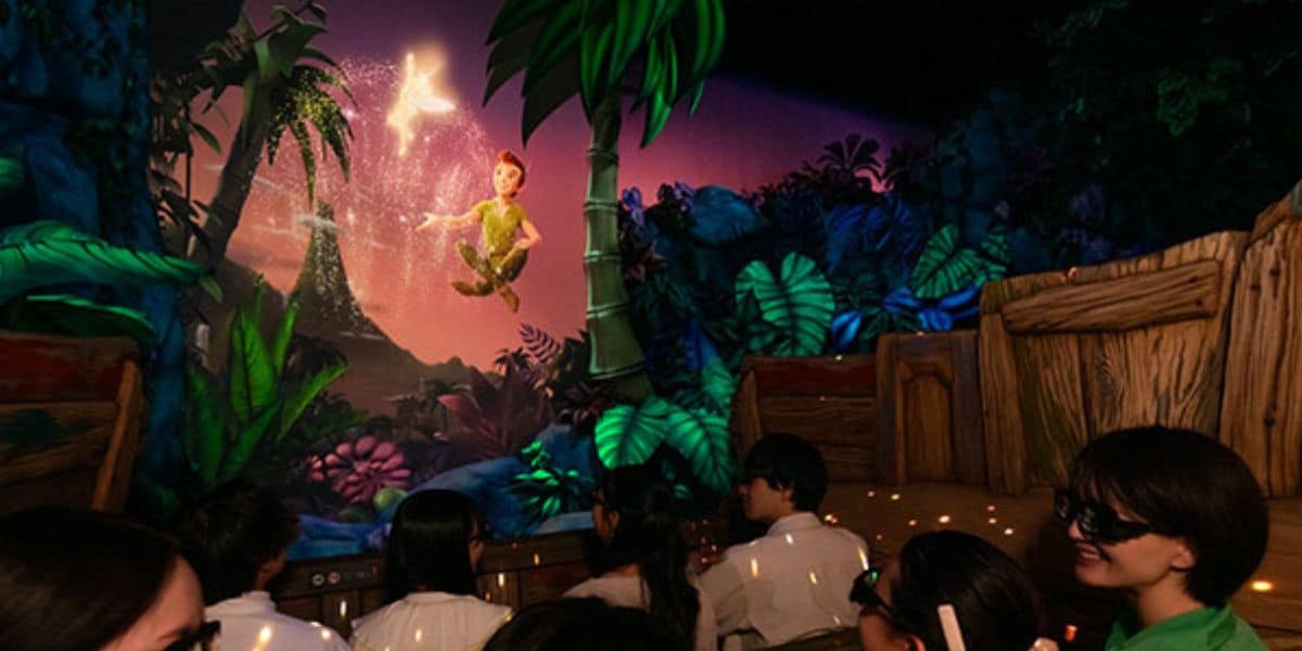 A group of people wearing 3D glasses sit in Disney's newest theme park ride, facing a vibrant, illuminated scene featuring a flying figure surrounded by sparkles and tropical foliage. The background depicts a colorful fantasy landscape with large trees and jungle plants; filming is banned here.