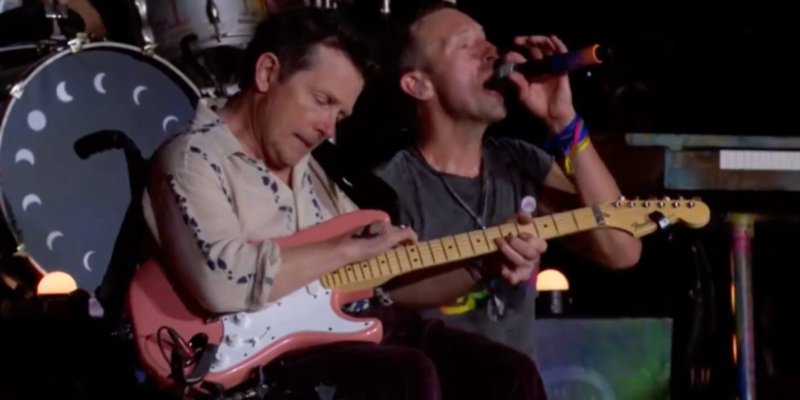 Two musicians are delivering a great performance on stage. One is playing an electric guitar, reminiscent of Michael J. Fox in Back to the Future, while the other passionately sings into a microphone, sporting a colorful striped wristband. In the background, a drum set with crescent moon designs can be seen as they sit close together.