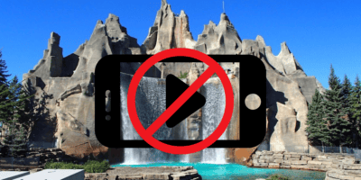 A picturesque scene of a mountain-like structure with waterfalls flowing into a pool is displayed. Centered in the foreground is an overlay of a mobile device with a red prohibition symbol, indicating "No phones," adhering to park bans on filming rides for YouTube.