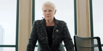 A stern-looking woman with short, gray hair stands leaning forward with her hands on a table. She is wearing a dark blazer over a black top and a brooch. She is in a well-lit room with large windows in the background.