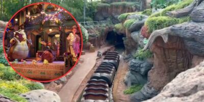 A rollercoaster ride in a theme park is depicted, featuring a winding track through rocky terrain with lush greenery. An inset image shows colorful animatronic dinosaurs in a lively, decorated setting. The scene appears vibrant and engaging, perfect for fans of Tiana's Bayou Adventure.