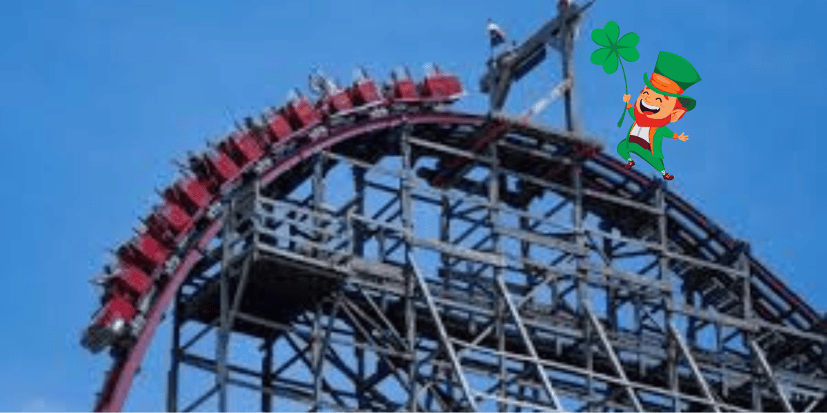 A red roller coaster climbs up a steep track structure against a clear blue sky. A whimsical cartoon leprechaun holding a four-leaf clover floats near the top right of the coaster, adding a playful element to the scene.