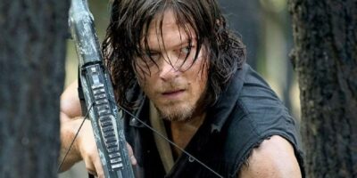 A person with long, dark hair and a serious expression holds a crossbow while crouching among trees. They are dressed in a dark, sleeveless shirt and appear to be in a forest setting, conveying a sense of alertness and readiness.