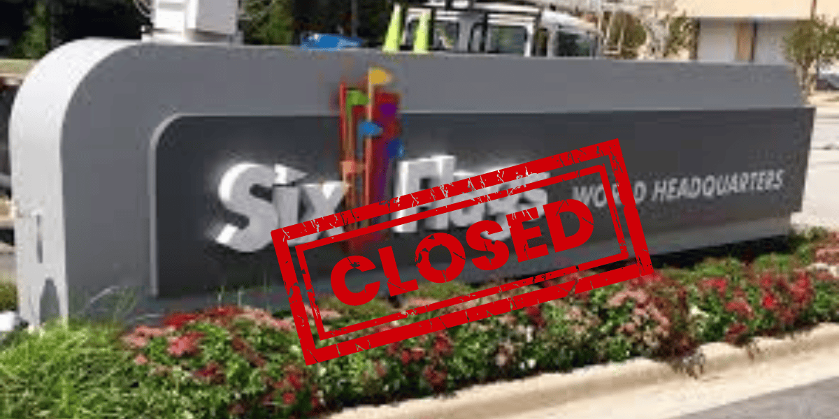 A sign indicating the corporate headquarters of Six Flags, with the words "WORLD HEADQUARTERS" visible. A bold red "CLOSED" stamp is overlaid on the image. The sign is surrounded by plants and shrubs, giving a shuttered look to the once-busy location.