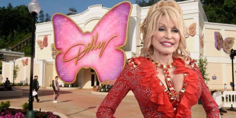 A cheerful woman with blonde hair wearing a red, jeweled outfit stands in front of a building decorated with large pink and beige butterfly designs. The largest pink butterfly in the background has the word “Dolly” written on it. People are walking around the area.