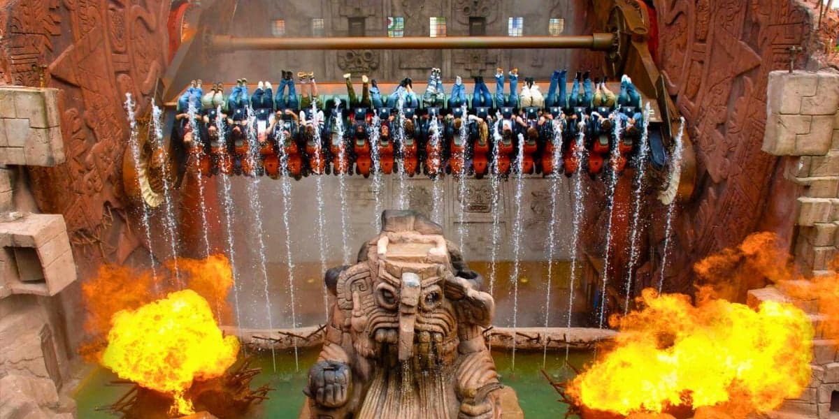 A theme park ride at Phantasialand with passengers seated in a suspended row rotates vertically above a stone statue resembling a mythical creature. Water pours down from the sides, and flames erupt in front of the statue, creating a dramatic scene.