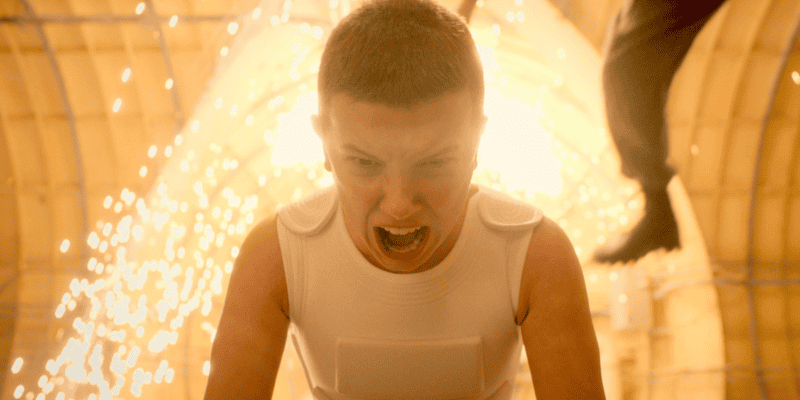 A person with a buzzcut and wearing a white sleeveless top is shown yelling intensely. Bright sparks and light erupt behind them in what appears to be a dramatic action scene. The background includes arched structures, suggesting an indoor setting.