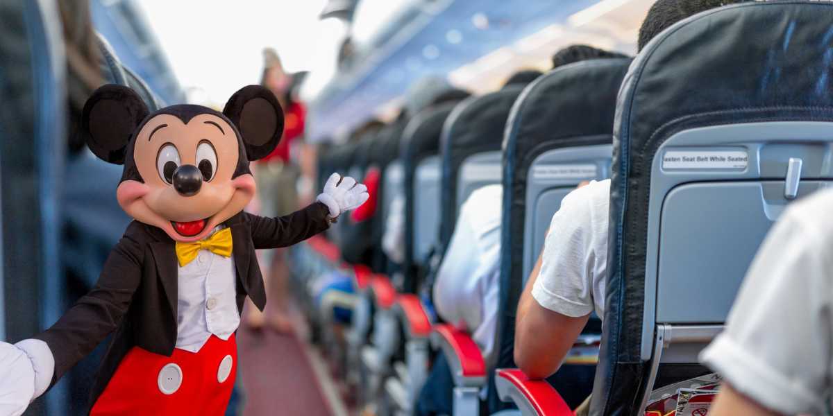 A person dressed as Mickey Mouse is waving and posing inside an airplane cabin on a Disney Flight. Passengers are seated in the airplane seats, and the aisle is visible in the background. The atmosphere appears lively and cheerful, adding a touch of magic to this popular airline experience.