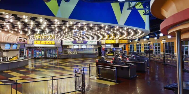 An empty and brightly lit cinema lobby features patterned tile flooring, multiple concession stands with illuminated signs advertising meals, and organized queues. The colorful ceiling with geometric shapes enhances the lively atmosphere, while the seating area is visible in the background.