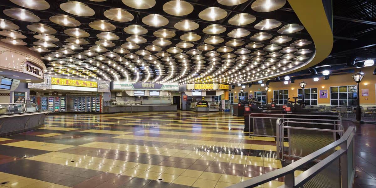 A brightly lit and spacious cafeteria with an array of round lights on the ceiling. The area features checkered tile flooring in shades of yellow, brown, and black. Several food counters and menus are visible, ready to serve thousands of meals daily, with tables and chairs positioned towards the right.
