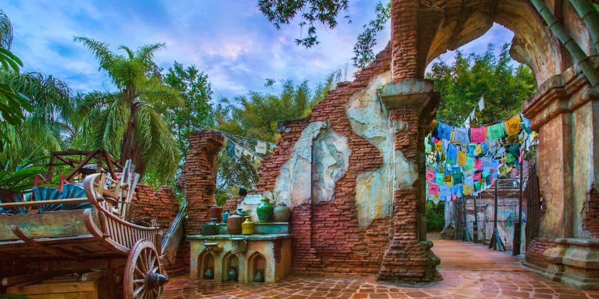 An outdoor scene featuring a rustic, partially crumbled brick wall with archways, vibrant colored prayer flags hanging above, large tropical plants in the background, and various pottery and wooden items on the ground. The sky is painted in shades of purple and blue.