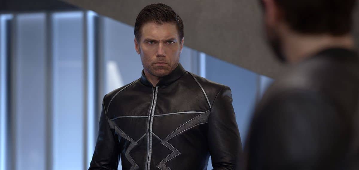 A man with short dark hair and a serious expression is wearing a black leather jacket with silver accents. He is standing indoors, facing slightly to his right with a blurred figure in the foreground on the right.