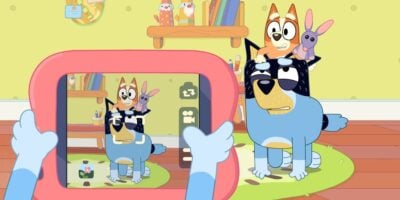 A scene from the animated series "Bluey" shows Bluey taking a photo of her dad, Bandit, who looks tired. Bluey is sitting on Bandit's back, holding a toy bunny, while another toy bunny rests on Bandit's head. The background features toys and a shelf adorned with more beloved items from Bluey's world.