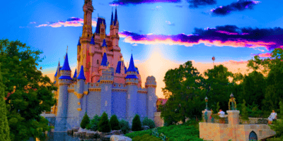 A picturesque view of Cinderella Castle with tall spires, set against a vibrant sunset sky with purple and pink clouds. The foreground features lush greenery and a pathway with visitors walking. The castle is surrounded by trees and a body of water.