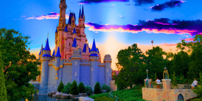 A picturesque view of Cinderella Castle with tall spires, set against a vibrant sunset sky with purple and pink clouds. The foreground features lush greenery and a pathway with visitors walking. The castle is surrounded by trees and a body of water.