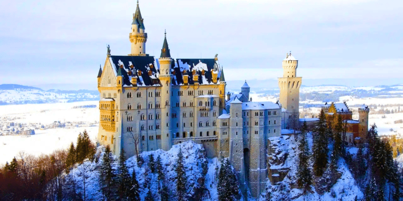 Neuschwanstein Castle in Bavaria, Germany, covered in snow. The picturesque castle, often compared to Cinderella Castle, sits majestically on a rugged hillside surrounded by snowy trees and a winter landscape, showcasing its tall towers, pointed spires, and intricate architectural details.