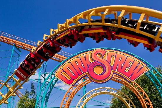 A colorful roller coaster named "Corkscrew" featuring a twisting yellow track with red cars full of passengers. The bright blue sky and vibrant support structures in orange and teal add to the lively atmosphere. Trees are visible at the bottom.