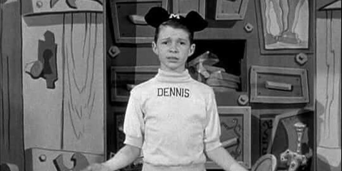 A young boy wearing Mickey Mouse ears with an "M" on them stands with his arms outstretched, looking into the camera. He is dressed in a white shirt that has the name "DENNIS" printed on it. The background features a cartoonish set design with various abstract shapes.