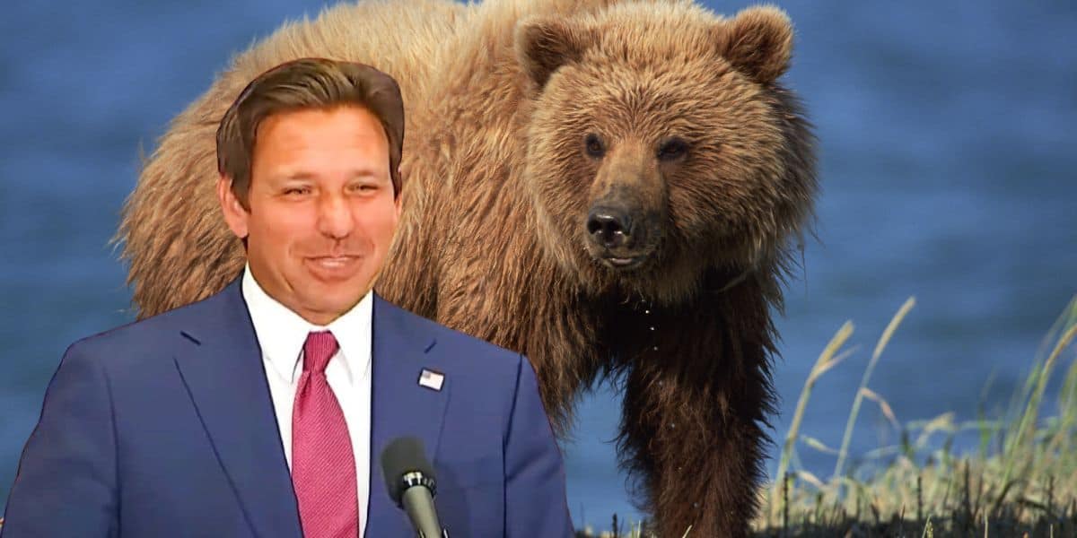 A man in a suit and red tie is standing and smiling, with his hand resting on a microphone. Behind him is the image of a brown bear near water and grass. The background and foreground images are blended together.