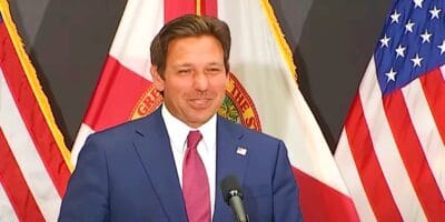 Ron DeSantis is standing at a podium, smiling. Behind him are the flags of the United States and Florida as Epic Universe opens.