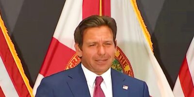 Ron DeSantis is standing in front of a backdrop featuring red and white flags. He has short brown hair and is speaking or making a gesture with his right hand.