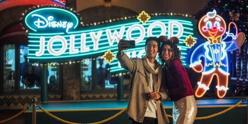 A smiling couple takes a selfie in front of a brightly lit Disney Jollywood Nights sign