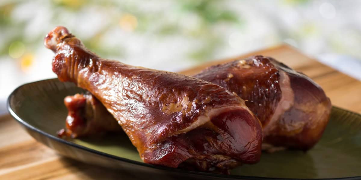 A close-up of two roasted turkey legs on a dark green plate. The meat appears tender and well-cooked, with a rich, glossy brown color. The plate is set on a wooden surface, and the background is softly blurred, emphasizing the focus on the turkey legs.