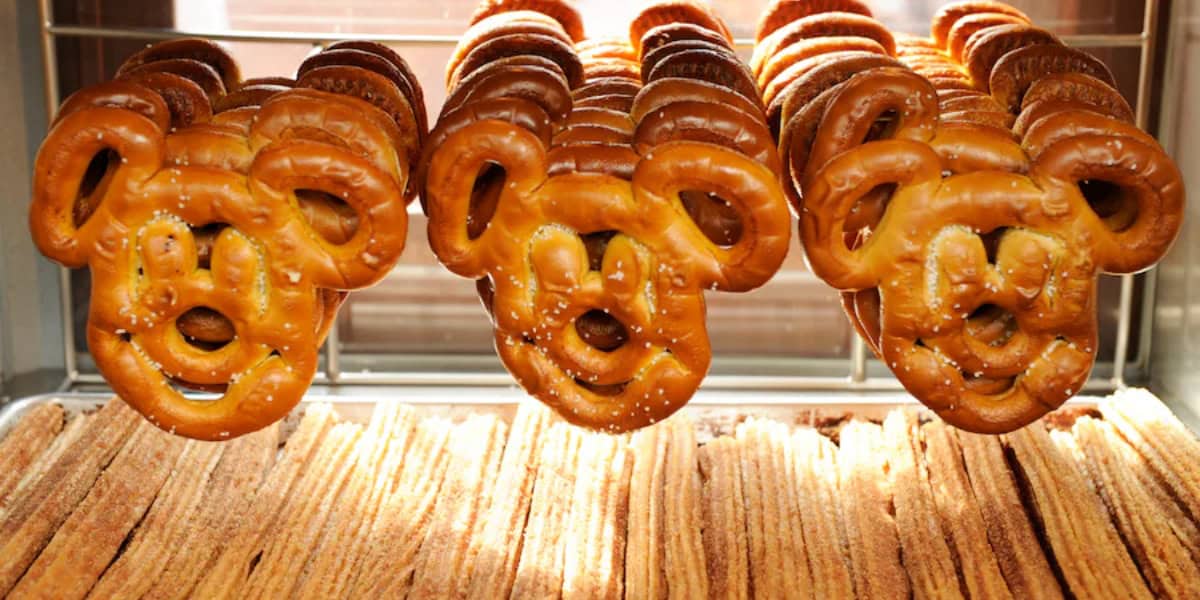 Three Mickey Mouse-shaped soft pretzels are displayed on a metal rack, with rows of golden churros underneath. The pretzels are a rich brown color with visible salt crystals, and the arrangement creates a pleasing display of popular snack foods.
