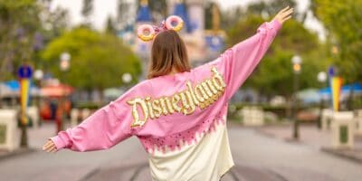 A person wearing a pink and white "Disneyland Resort" jacket and donut-themed Mickey ears stands with arms outstretched on an empty Main Street, USA, facing towards Sleeping Beauty Castle at Disneyland. The scene is vibrant, with greenery and buildings in the background.