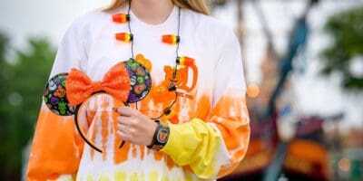 A person wearing a white and orange ombre sweatshirt, a necklace with candy corn lights, and a black wristband with Halloween designs. They are holding a headband with black mouse ears decorated in orange and green Halloween patterns and an orange bow.