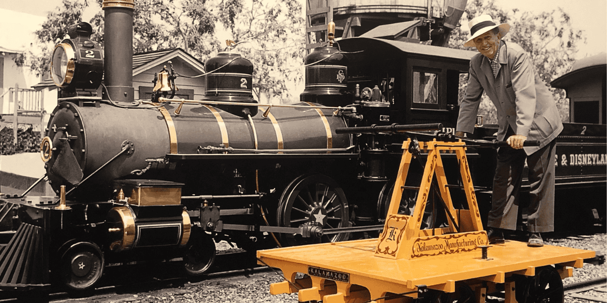 A man wearing a hat and glasses stands on a yellow handcar next to a black steam locomotive with the number 2 on it, reminiscent of Disneyland's vintage charm. Trees are visible in the background, enhancing the nostalgic ambiance.