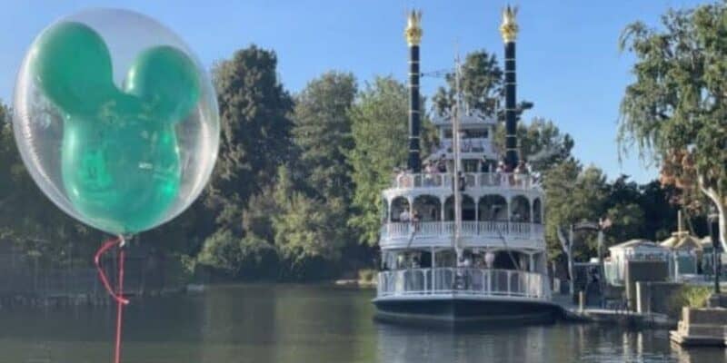 A large, green Mickey Mouse-shaped balloon floats in the foreground on the left side. In the background is the Rivers of America at Disneyland Park, with the Mark Twain riverboat.