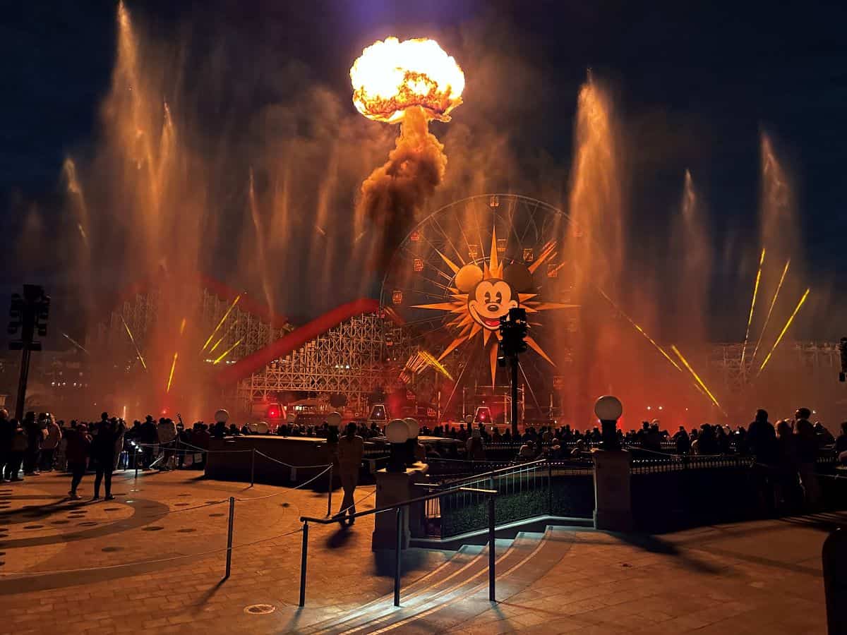 The Incredicoaster and Pixar Pal-A-Round in Disney California Adventure's Pixar Pier during a presentation of World of Color — ONE at the park. The scene shows a massive fire column rising in the middle of the lagoon, a culminating moment of the nighttime spectacular that often leaves massive rings of black smoke floating above Disneyland Resort
