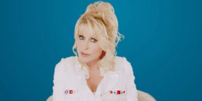 A blonde woman with styled hair stands against a solid blue background. She is wearing a white jacket with nautical flag designs on it and a white top underneath. She has red earrings, subtle makeup, and is gazing directly into the camera.