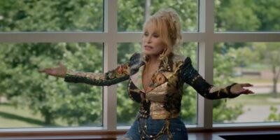 Dolly Parton in a colorful, patterned jacket and blue jeans, gesturing with both arms wide open in front of a large window with green trees visible outside.